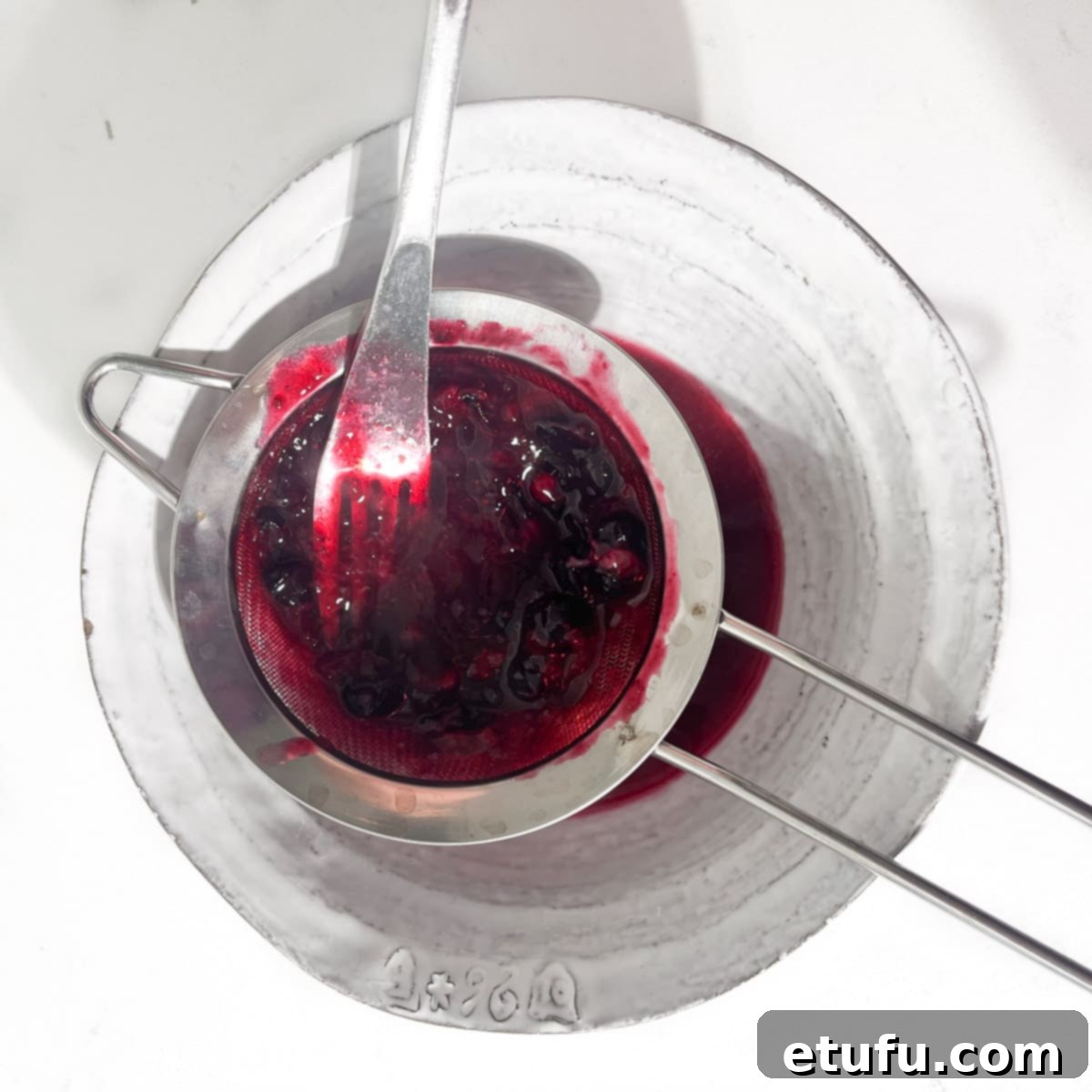 Blueberries being squashed with a fork through a sieve into a white bowl below.