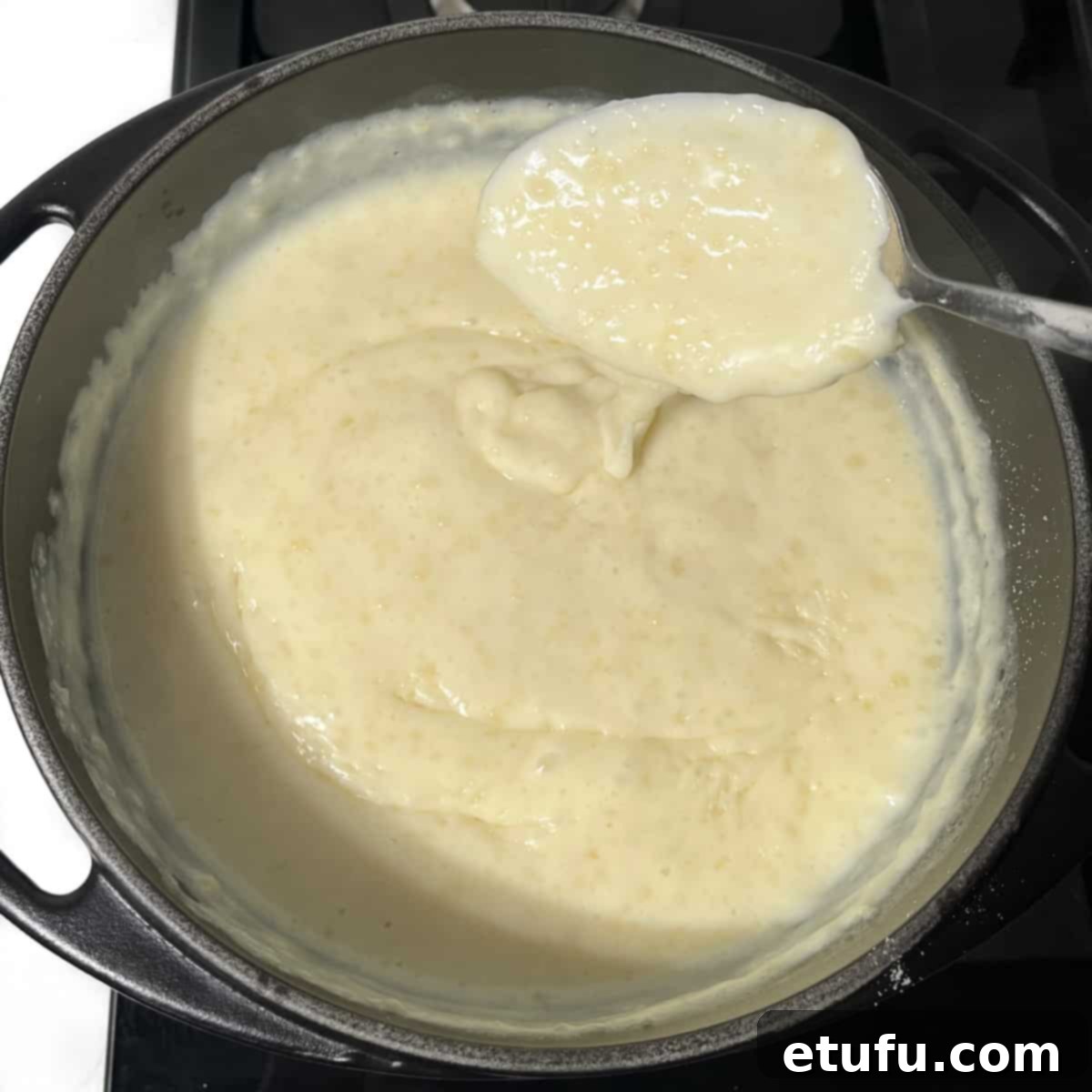 A large spoonful of creamy Melkkos held above a black saucepan, showcasing its rich texture and warmth, ready for serving.