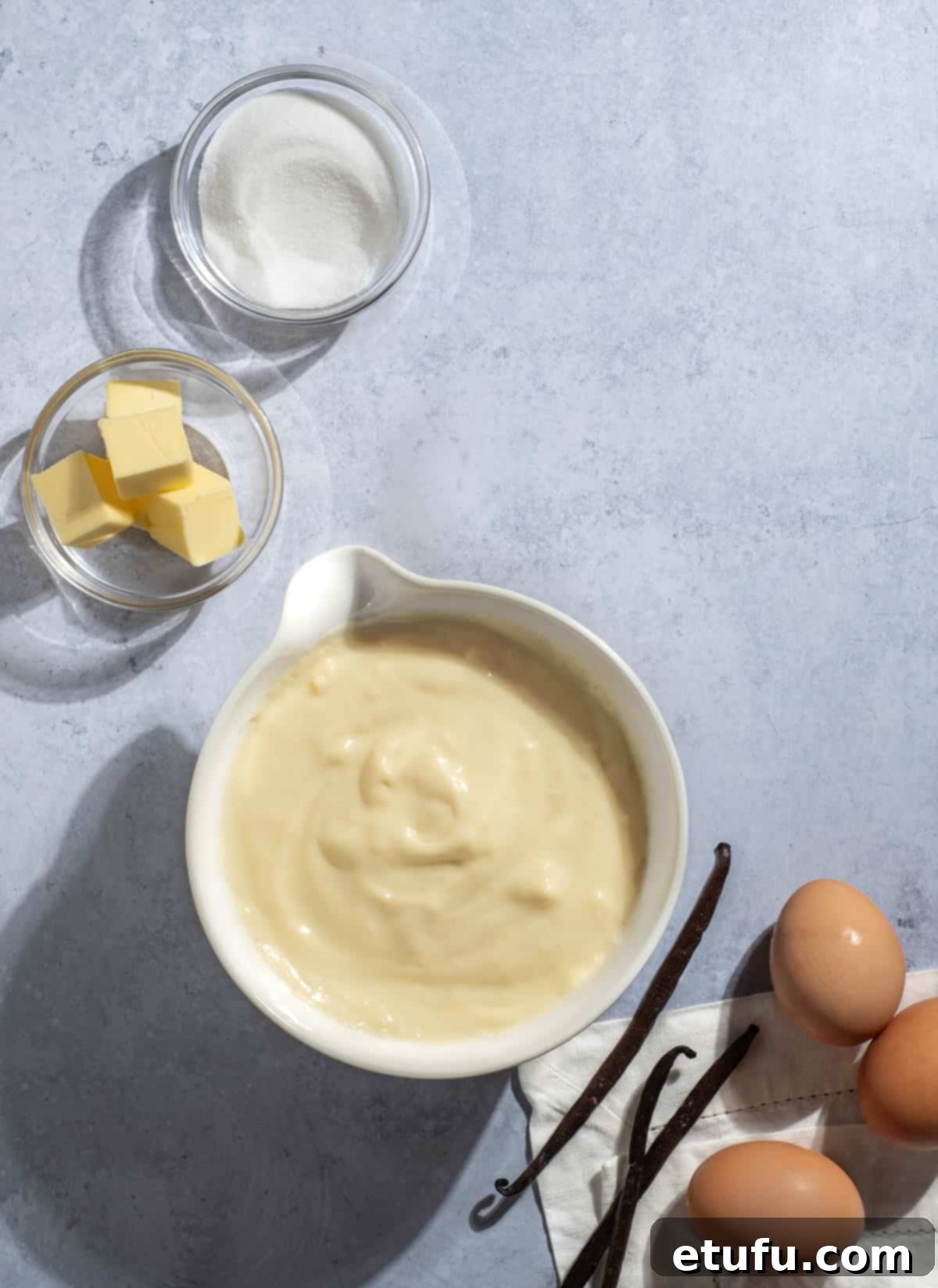 A bowl of Crème Pâtissière on a grey background surrounded by eggs, vanilla pods, butter and sugar.