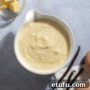 A bowl of Crème Pâtissière on a grey background surrounded by eggs, vanilla pods, butter and sugar.