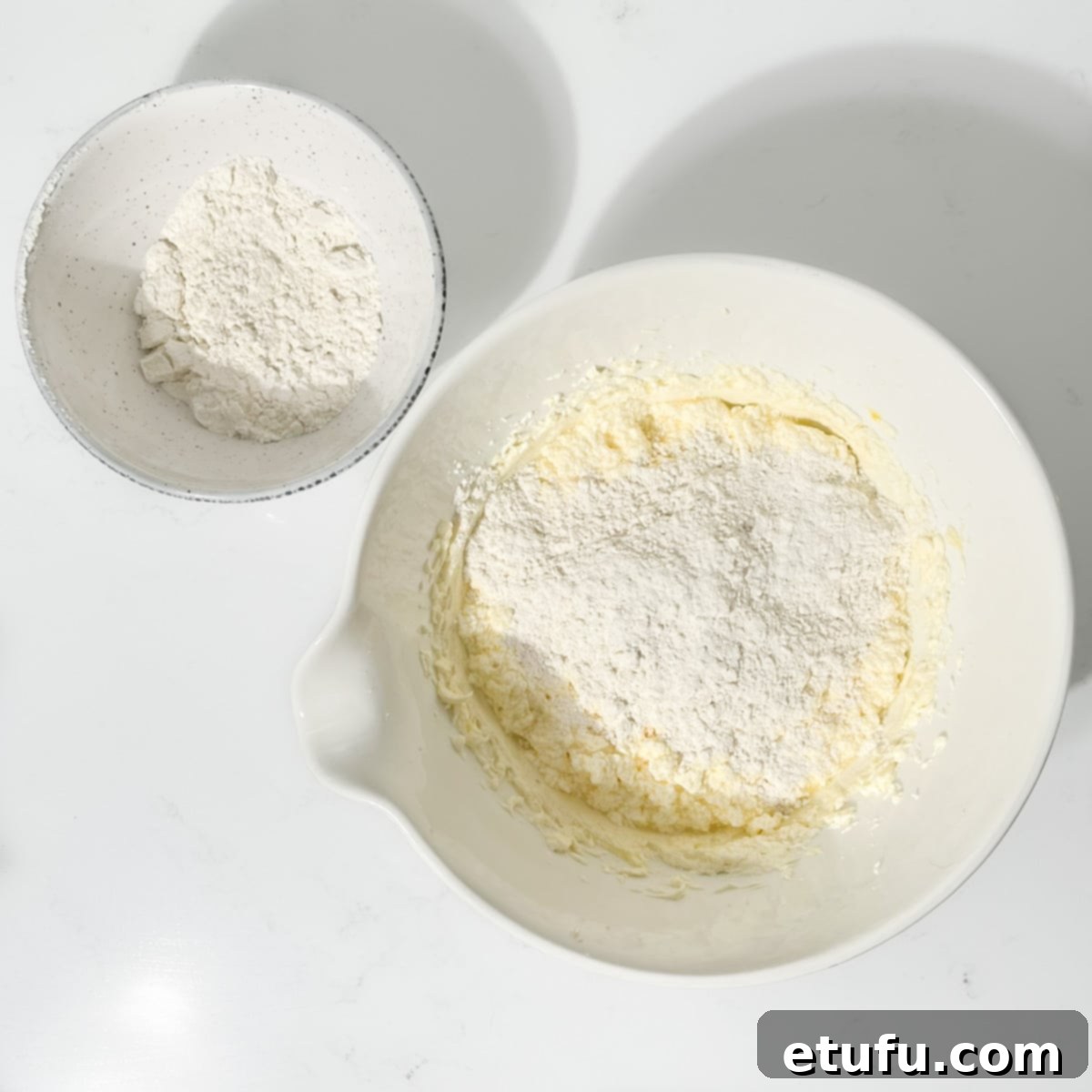 Adding half the flour to the batter in a large white bowl.