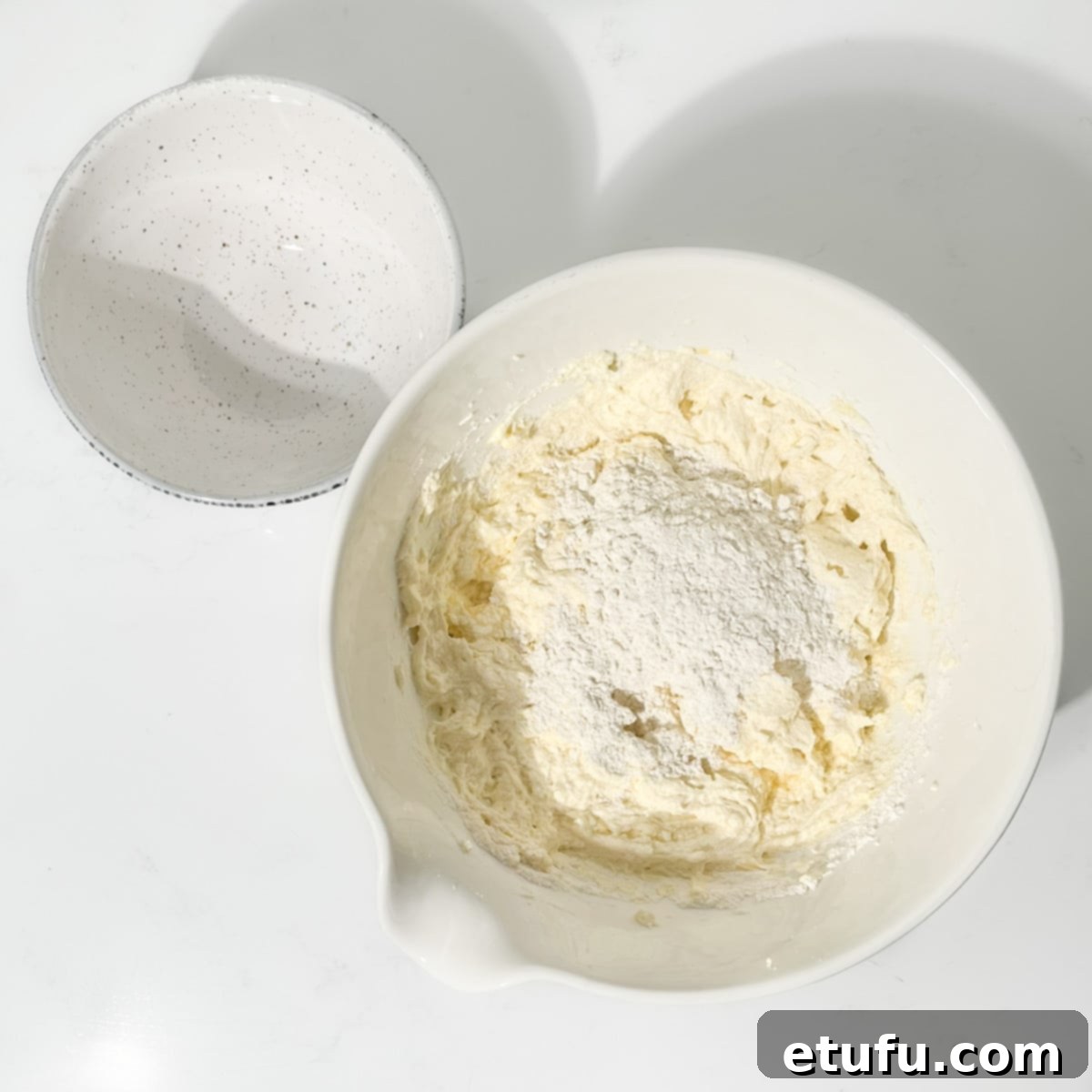 Adding the remaining flour to the batter in a large white bowl.