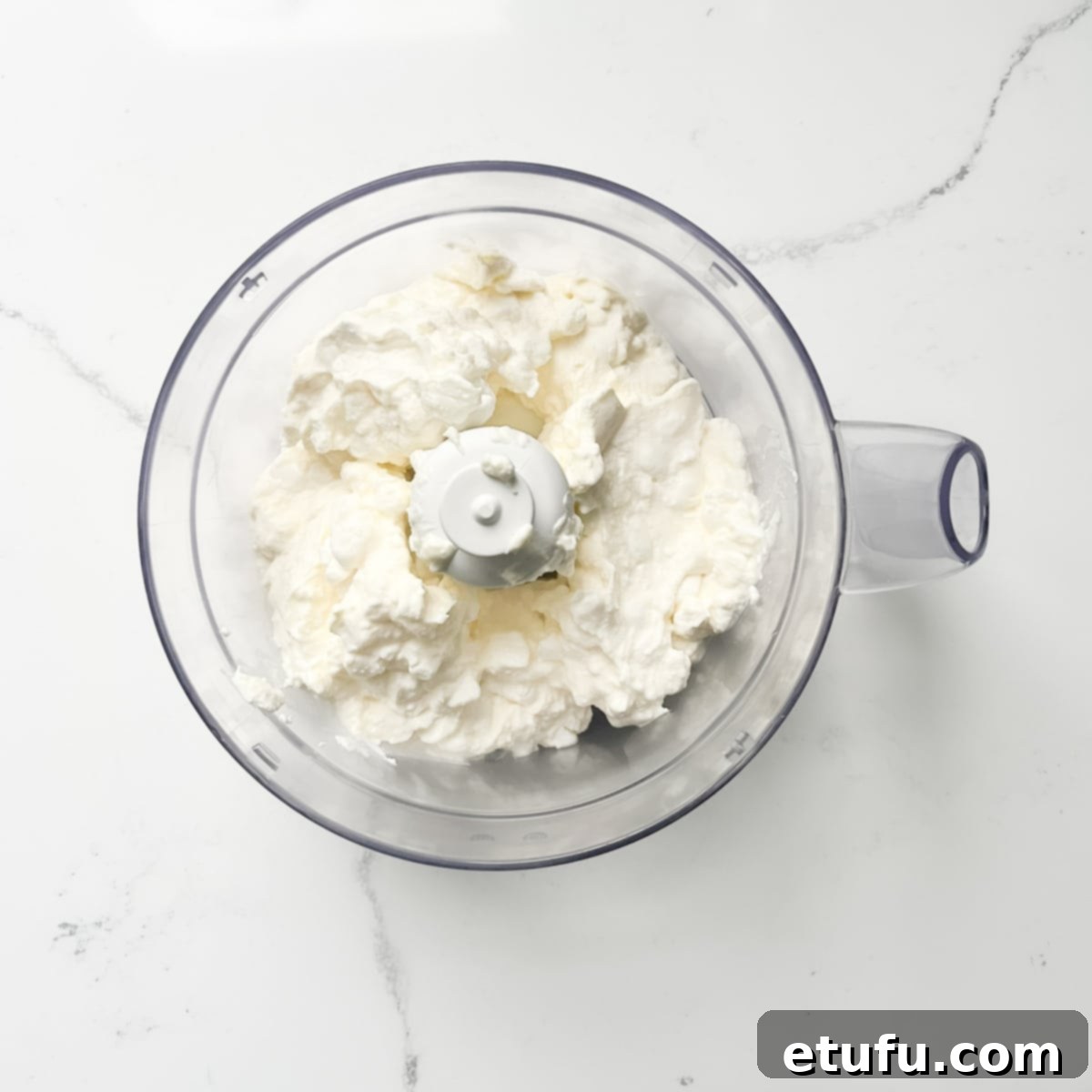 Crumbly cottage cheese being added to a food processor for whipping.
