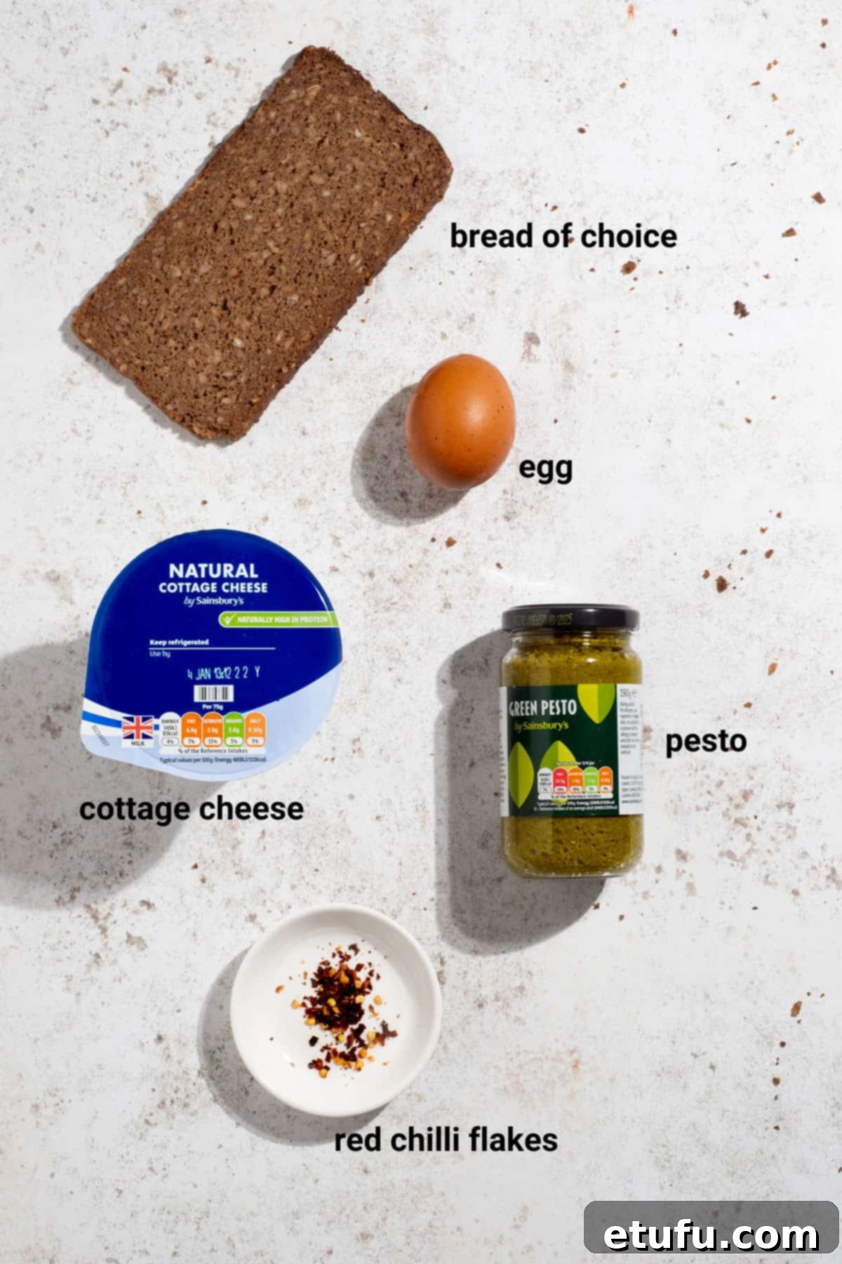 Ingredients laid out for fried egg and pesto cottage cheese toast, including an egg, pesto, chili flakes, and bread.