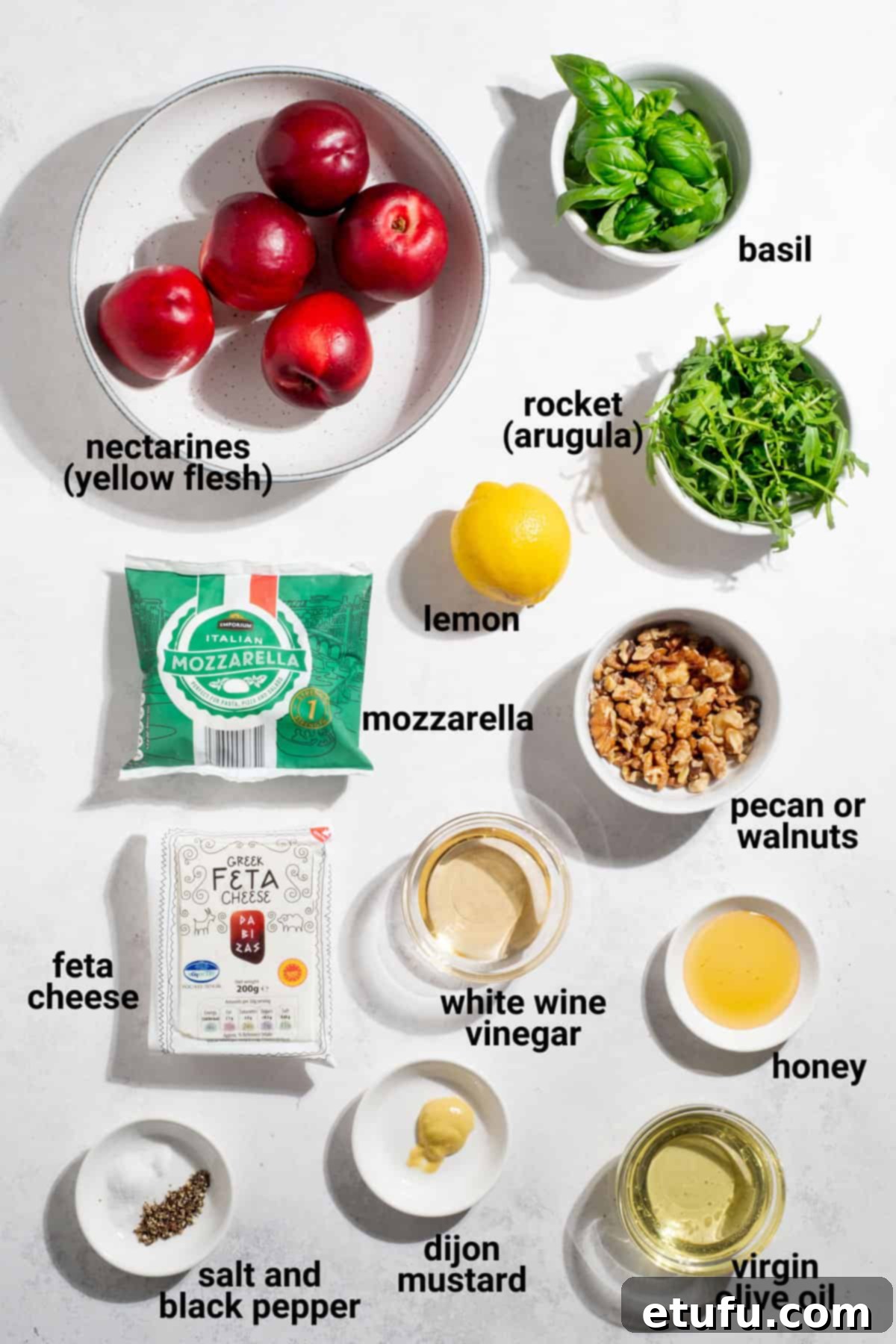 Ingredients for a fresh nectarine salad arranged neatly on a white surface, including vibrant sliced nectarines, a ball of mozzarella, crumbled feta, fresh basil leaves, and a small bowl with dressing components.