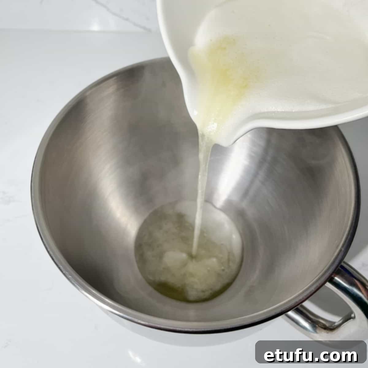 The warm egg and sugar mixture being poured into the bowl of a stand mixer, ready for whipping.