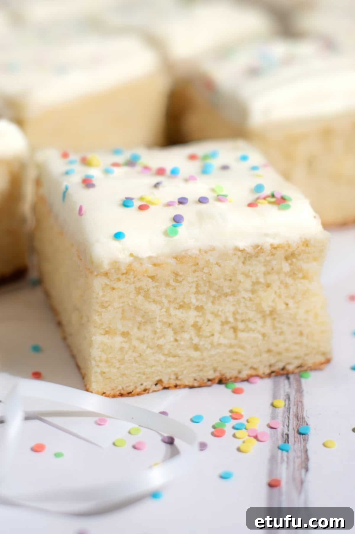 One generous slice of vanilla tray bake, topped with sprinkles and vanilla buttercream, on a white plate.