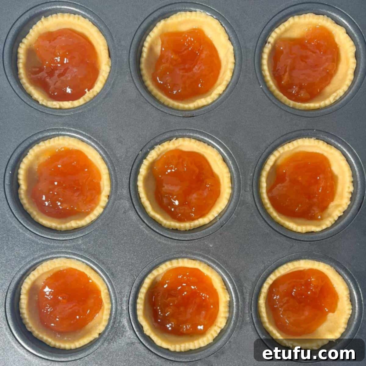 Apricot jam being spooned into the pastry cases in a muffin tray, ready for the meringue topping.