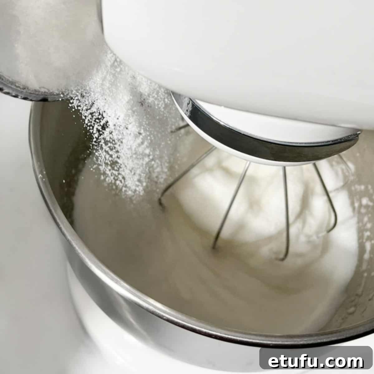 Granulated sugar slowly being added to the stiffly whipped meringue in a stand mixer, creating a glossy texture.