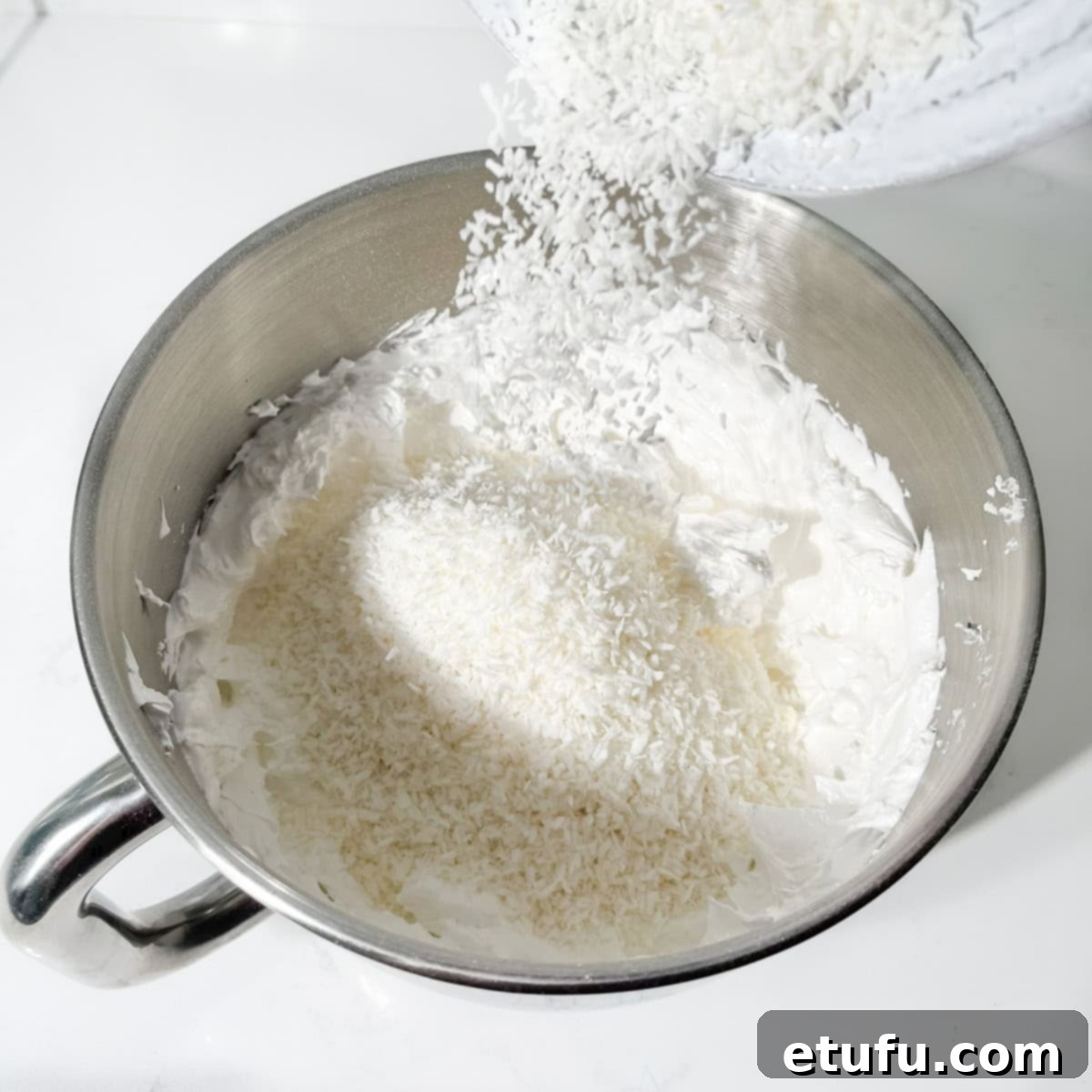 Desiccated coconut being added to the finished meringue mixture in a stand mixer bowl.
