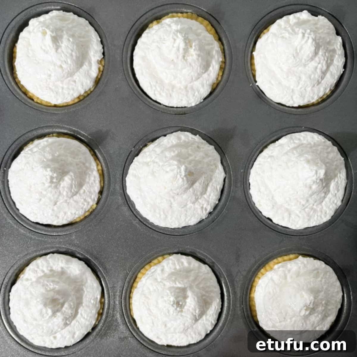 The coconut meringue topping being spooned onto the apricot jam-filled Hertzoggies in the muffin tray.