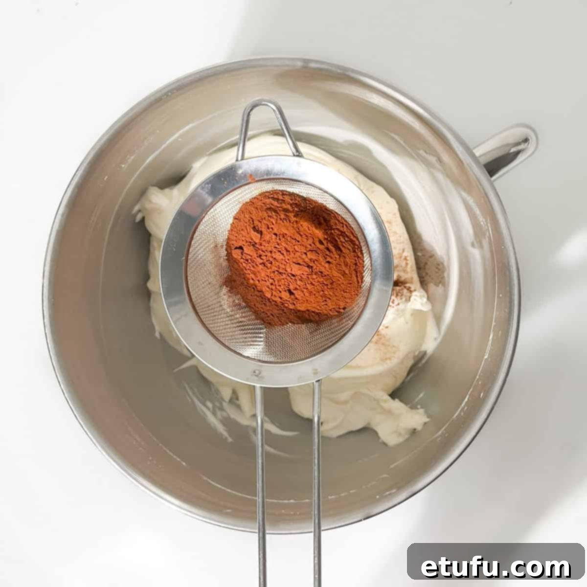 Sifting cocoa powder onto the cream cheese in the bowl of a stand mixer.