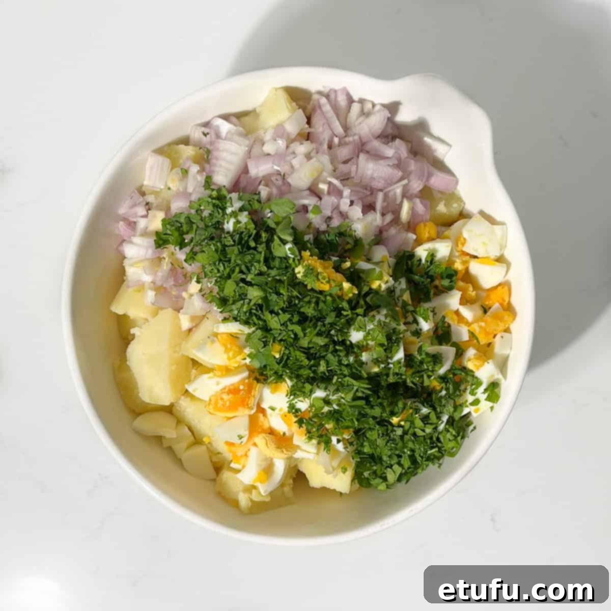 Adding the herbs, boiled eggs and onion to the sliced potato in a large white bowl.