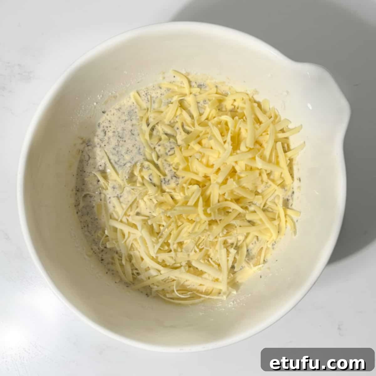Folding grated cheddar and Gruyère cheese into the quiche mixture in a large white bowl.