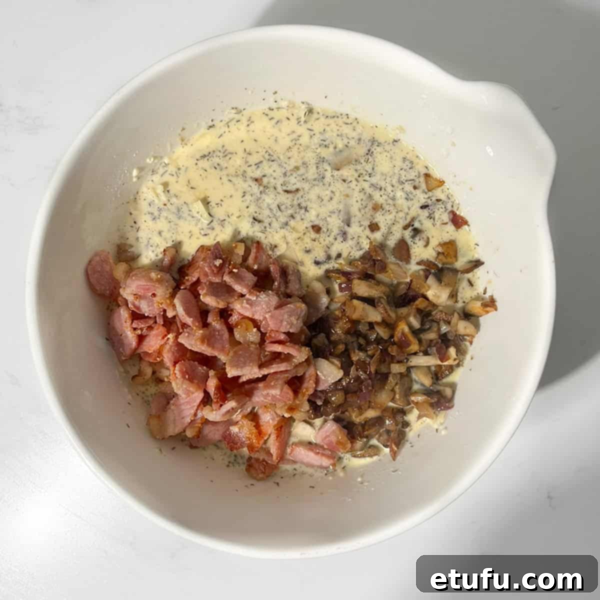 Adding cooked bacon and mushrooms to the creamy quiche batter in a large white mixing bowl.