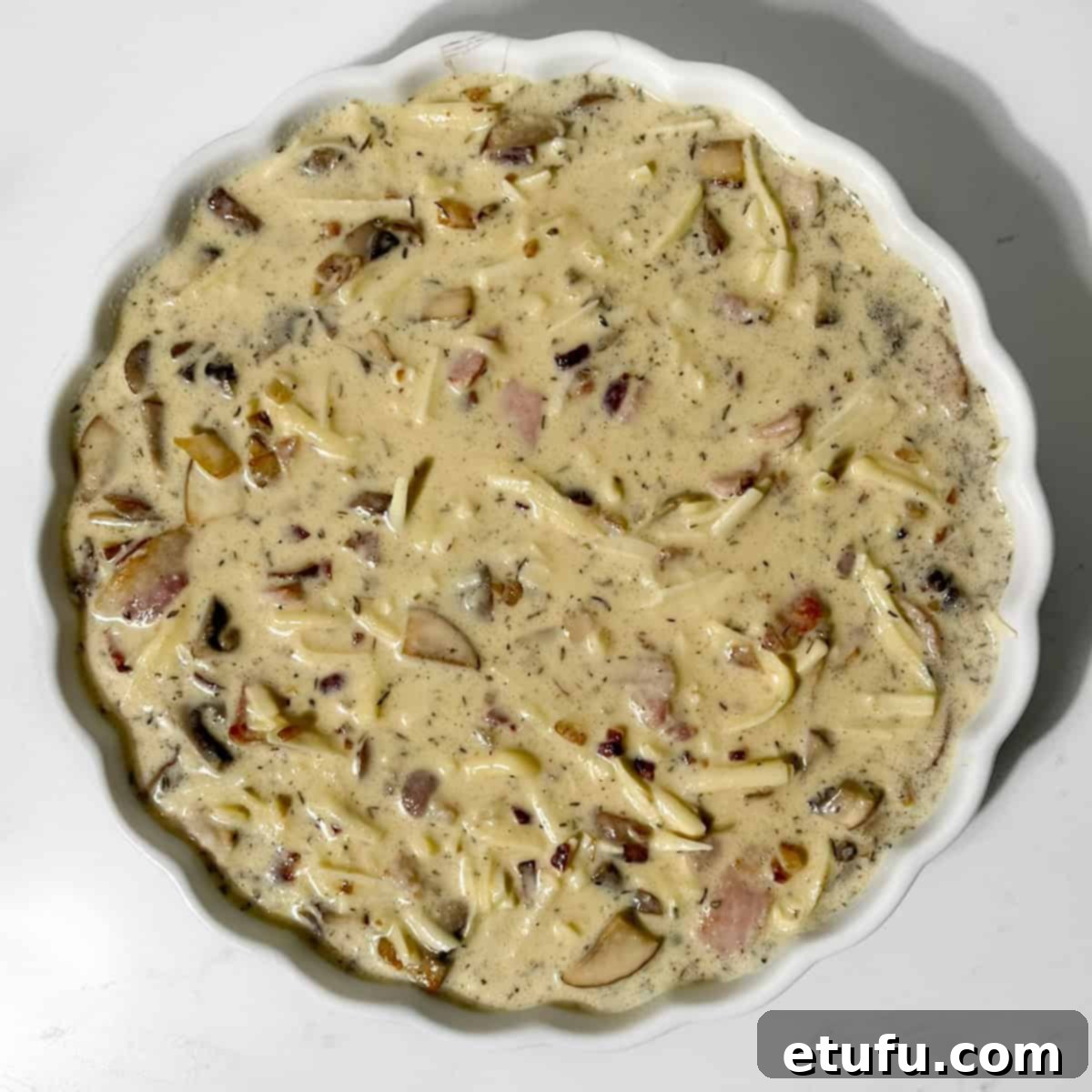 The quiche filling, with visible bacon and mushrooms, poured into a white tart dish, ready for baking.