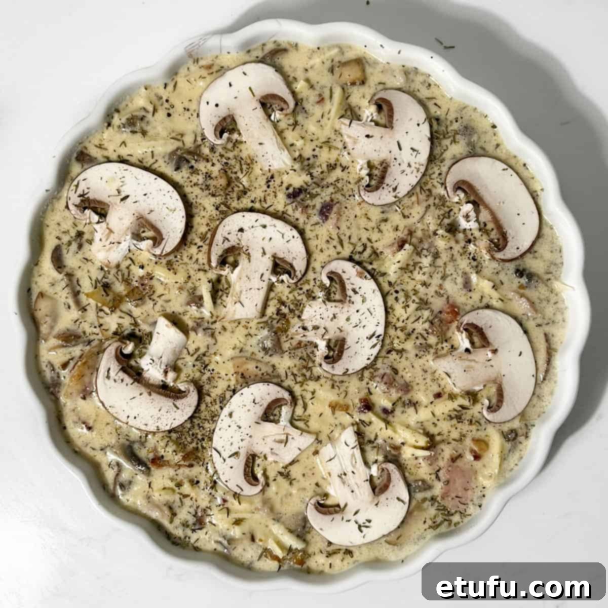 Unbaked quiche in a white tart dish, topped with additional mushroom slices, thyme, and black pepper for garnish.