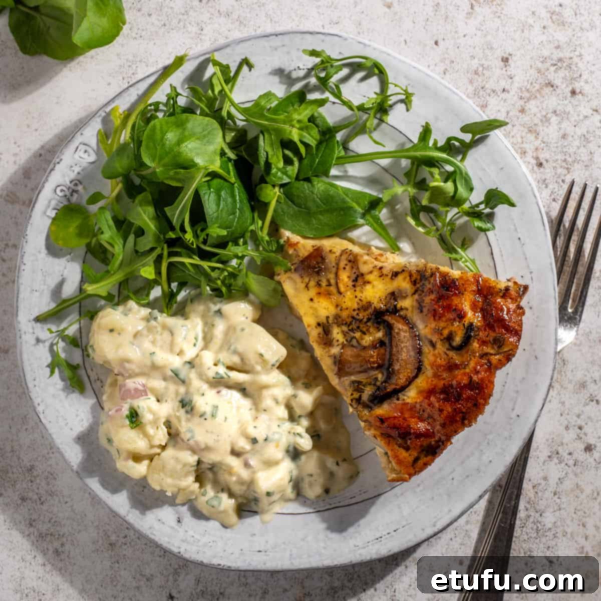 A slice of crustless bacon and mushroom quiche on a plate, accompanied by a vibrant green salad and creamy potato salad.