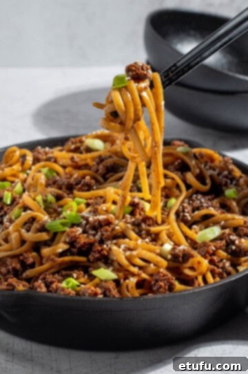 Mongolian ground beef noodles served in a large skillet, garnished with fresh green onions and toasted sesame seeds, with chopsticks poised to pick up a mouthful.
