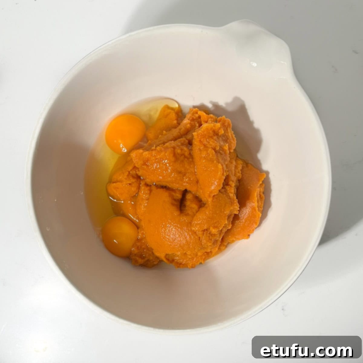 Two large eggs being cracked into a bowl of smooth pumpkin puree, the base for the pumpkin fritter batter, ready to be mixed.