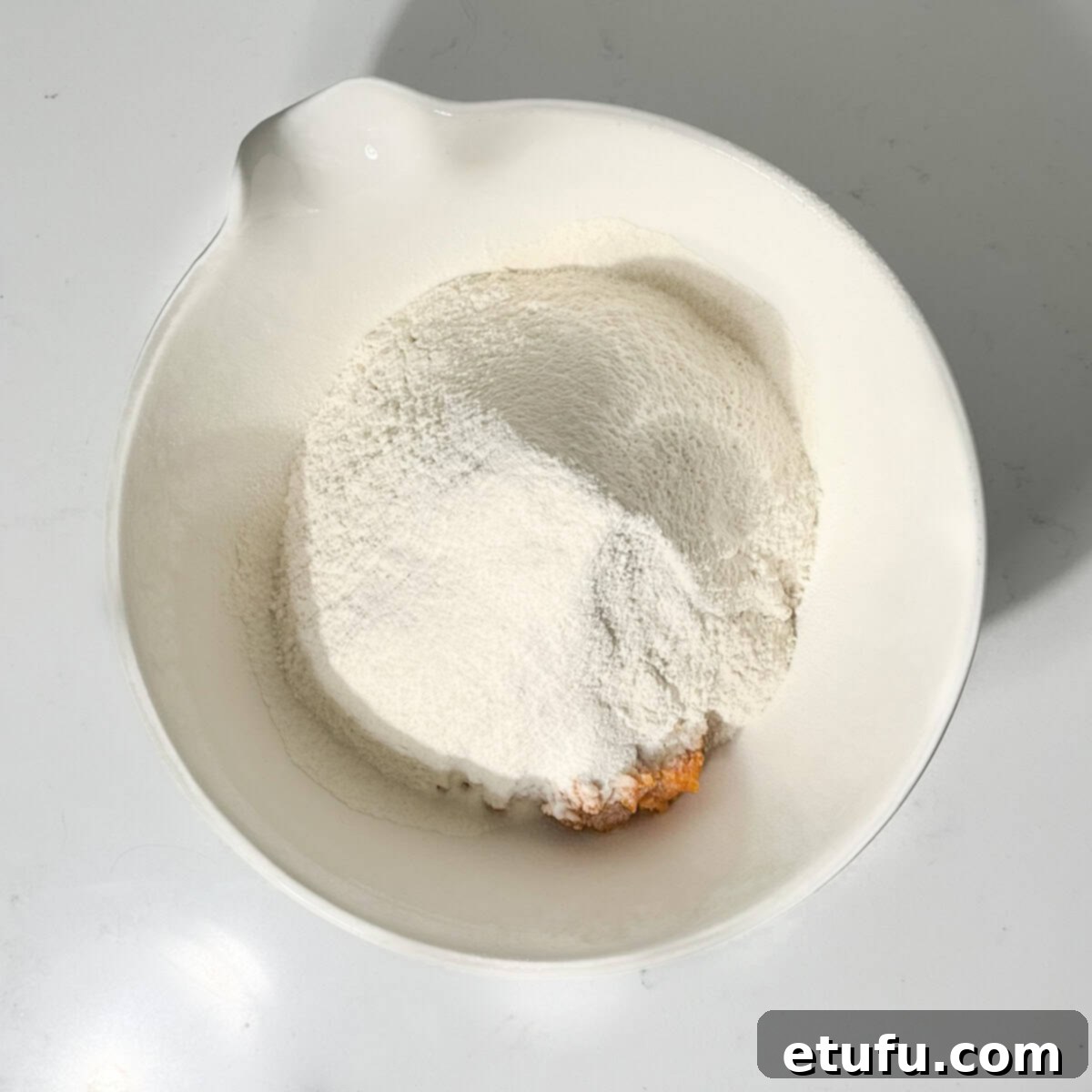 Sifting flour, baking powder, and salt directly into the pumpkin and egg mixture in a large white bowl, preparing for the pumpkin fritter batter.