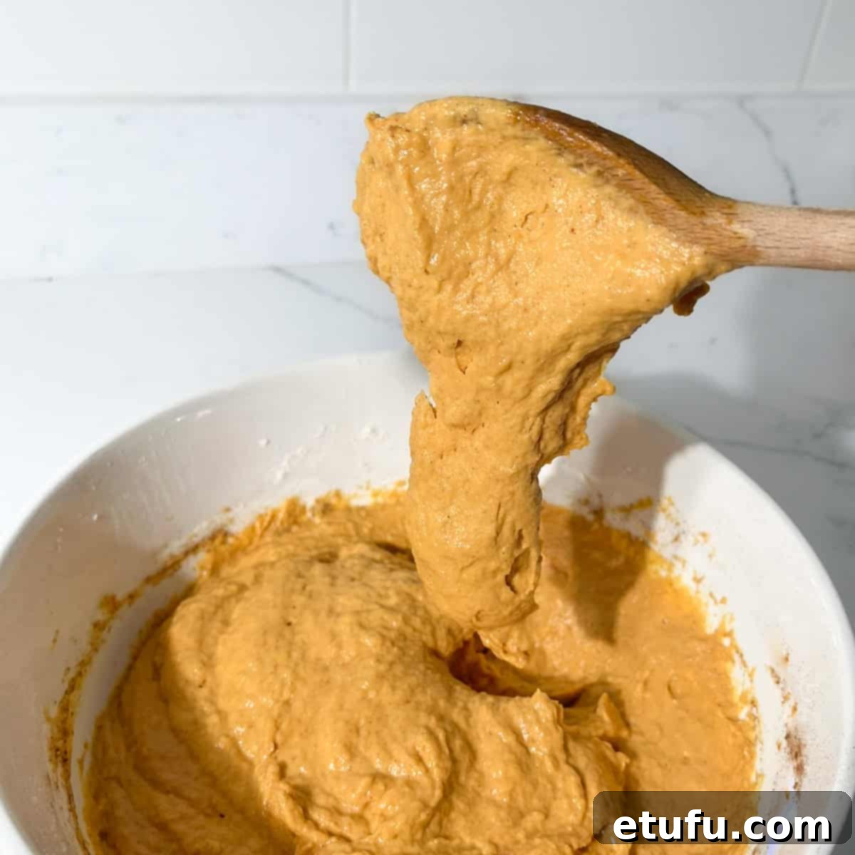 A wooden spoon slowly releasing a dollop of pumpkin fritter batter, demonstrating its thick yet soft consistency, holding its shape without being stiff.