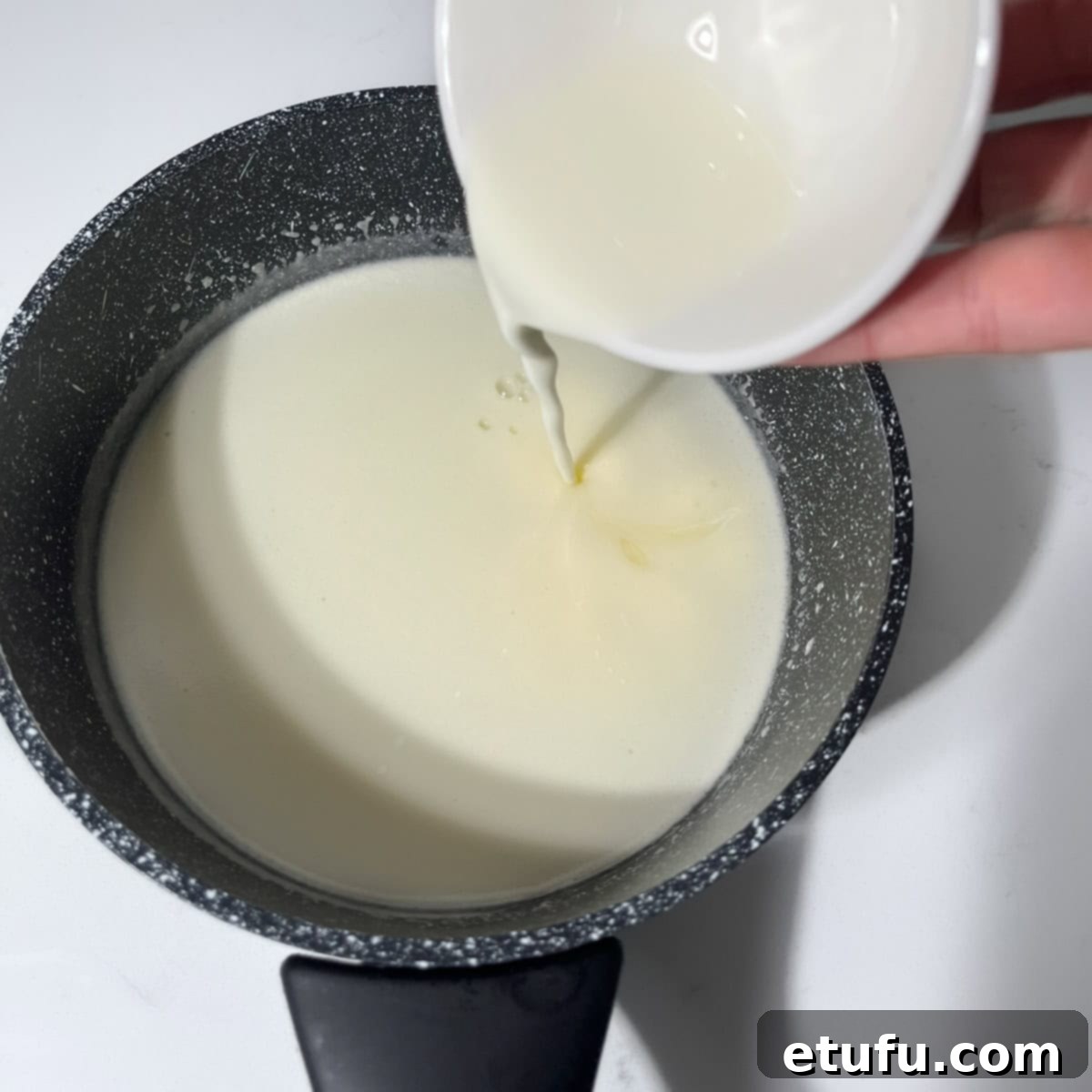 The cornstarch slurry is carefully poured into the simmering cream and sugar mixture in a small black saucepan, while stirring continuously to prevent lumps.