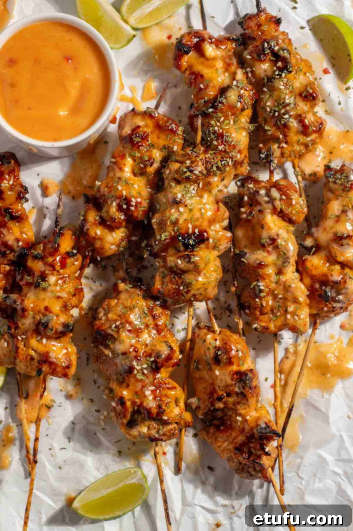Golden brown bang bang chicken skewers arranged neatly on a white background, accompanied by a small pot of creamy bang bang sauce and vibrant lime wedges.