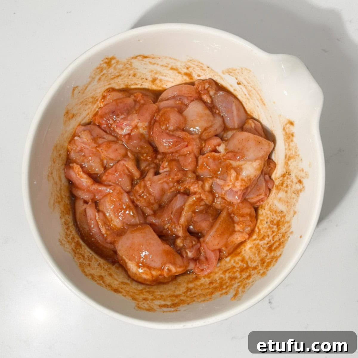 Cubed chicken pieces being stirred into a rich marinade in a large white bowl, ensuring every piece is coated.