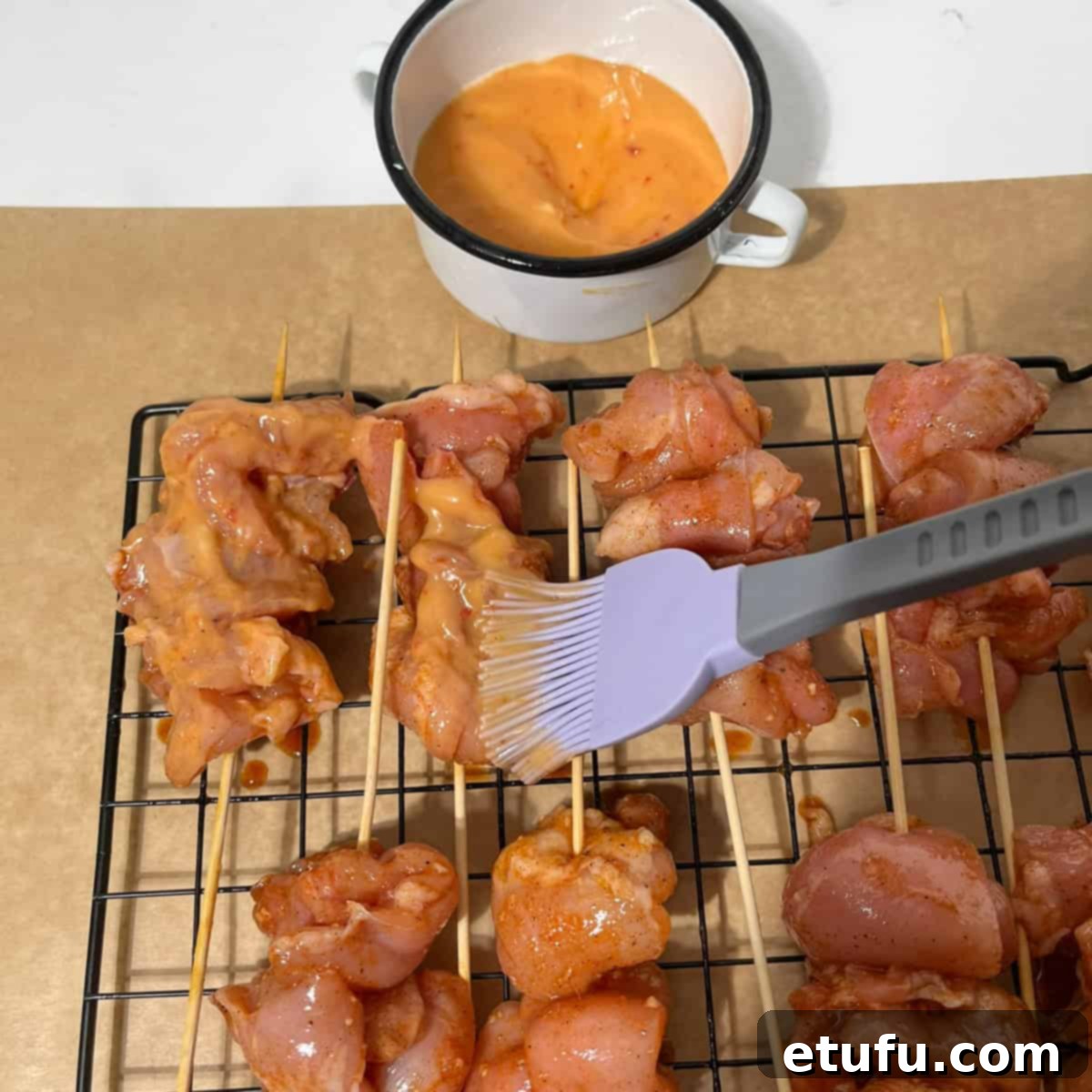 A brush applying a generous layer of bang bang sauce onto the raw chicken skewers arranged on a wire rack.