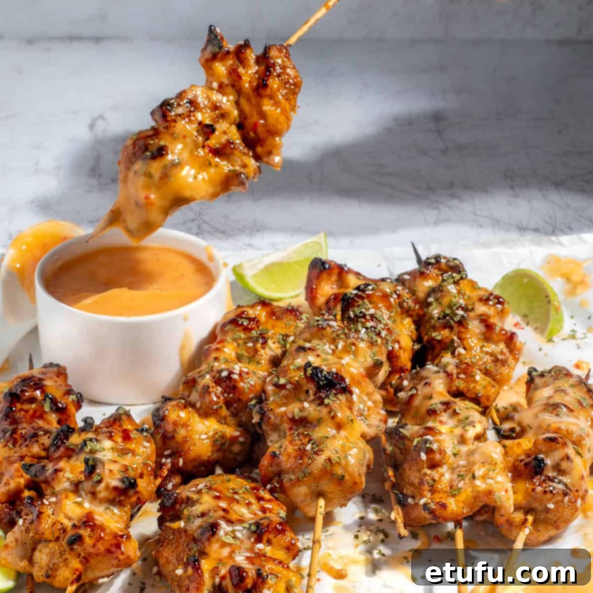 Appetizing bang bang chicken skewers presented on a pristine white background, accompanied by a small bowl of the signature sauce and fresh lime wedges.