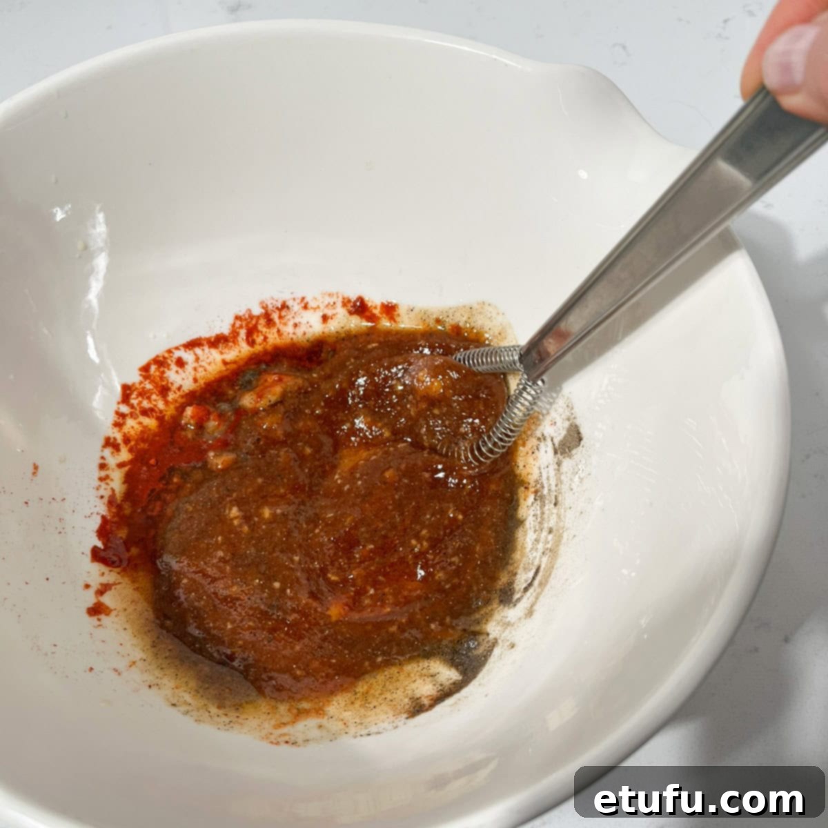 A hand whisking together marinade ingredients in a large white mixing bowl, showing the liquids and spices combining smoothly.